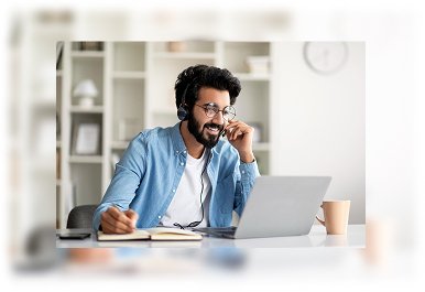 Man wearing headset working on a laptop while discussing project requirements in a modern office setting.
