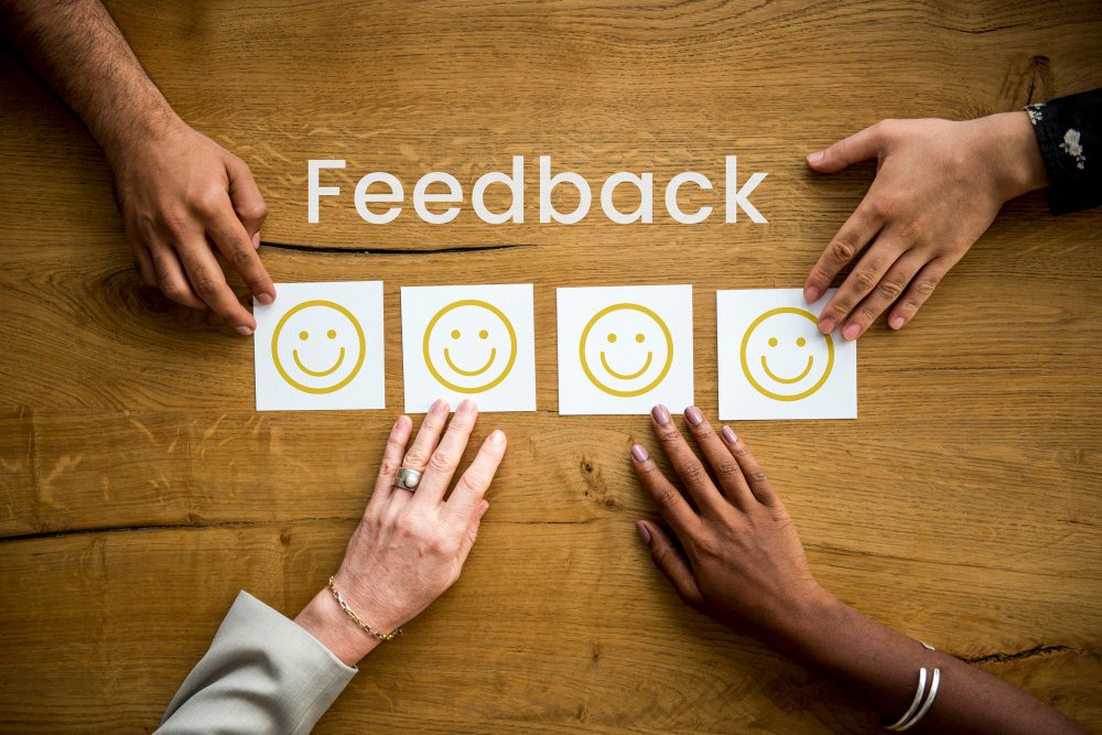 Hands placing smiley feedback cards on a wooden table under the word “Feedback.”