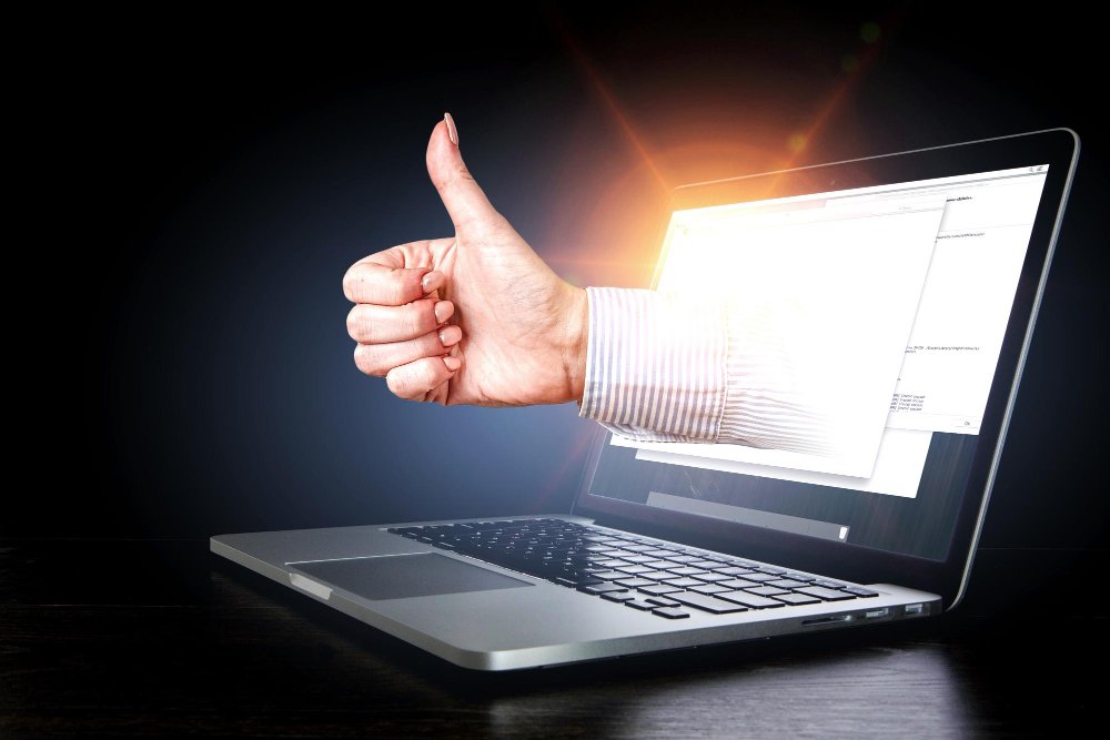 Hand giving thumbs up emerging from a laptop screen symbolizing approval and project launch.